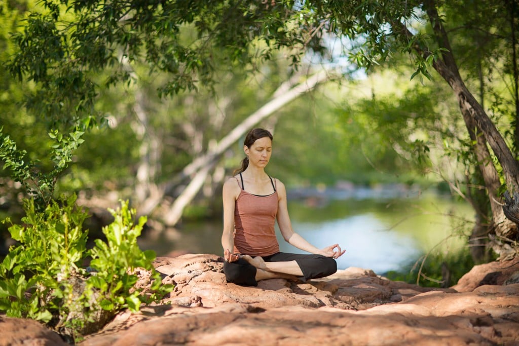 A woman practicing yoga in the position of lotus beside a creek under an archway of trees