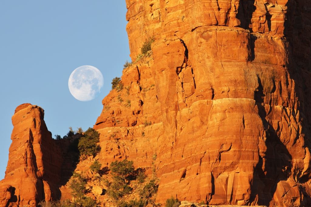 Full moon descends behind red rock desert butte as morning sun rises.