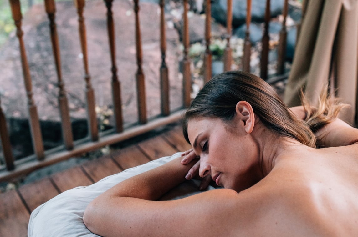 Woman lies face down on outdoor massage table with railing and rocks in background