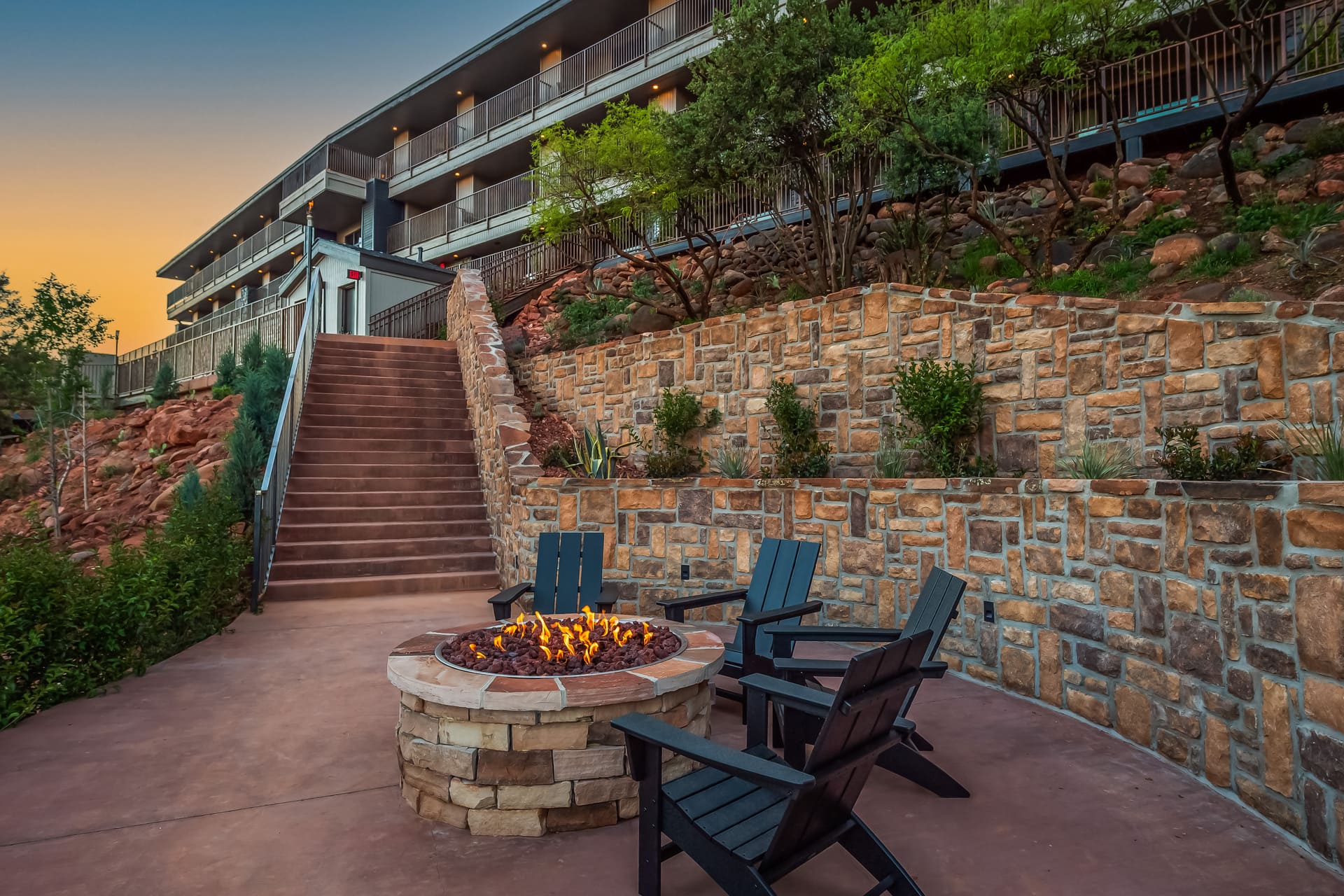 A fire pit with chairs in front of a stone wall and staircase leading to a multi-level building with balconies.