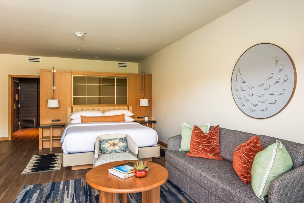 A hotel suite with a large bed, gray sofa, wooden coffee table, and decorative artwork.