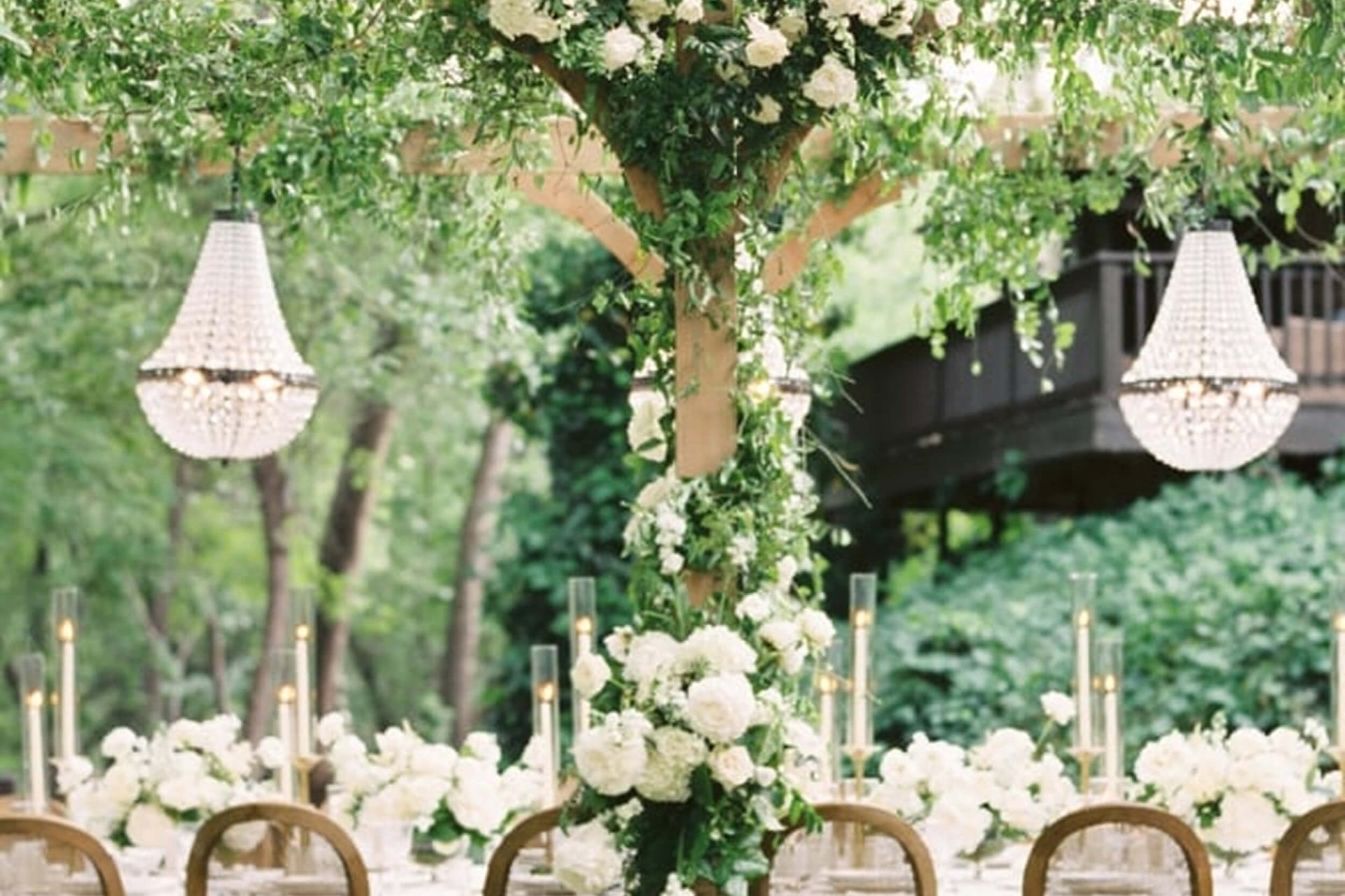 An elegant outdoor dining table under a wooden pergola, heavily decorated with white flowers and crystal chandeliers.