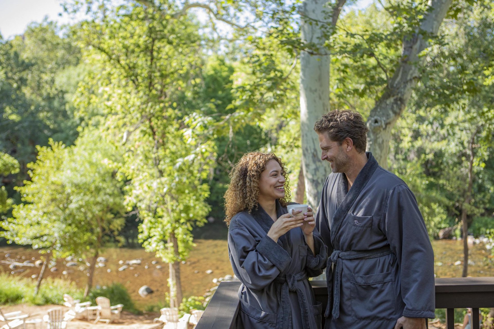 A smiling couple in gray bathrobes enjoys coffee on a private deck overlooking the lush trees and Oak Creek.