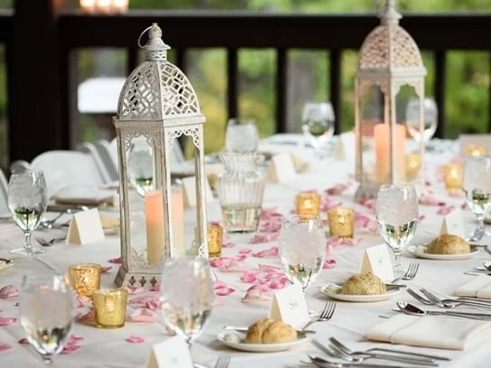 A long dining table set for a wedding, with white lanterns, wine glasses, and pink flower petals scattered on top.