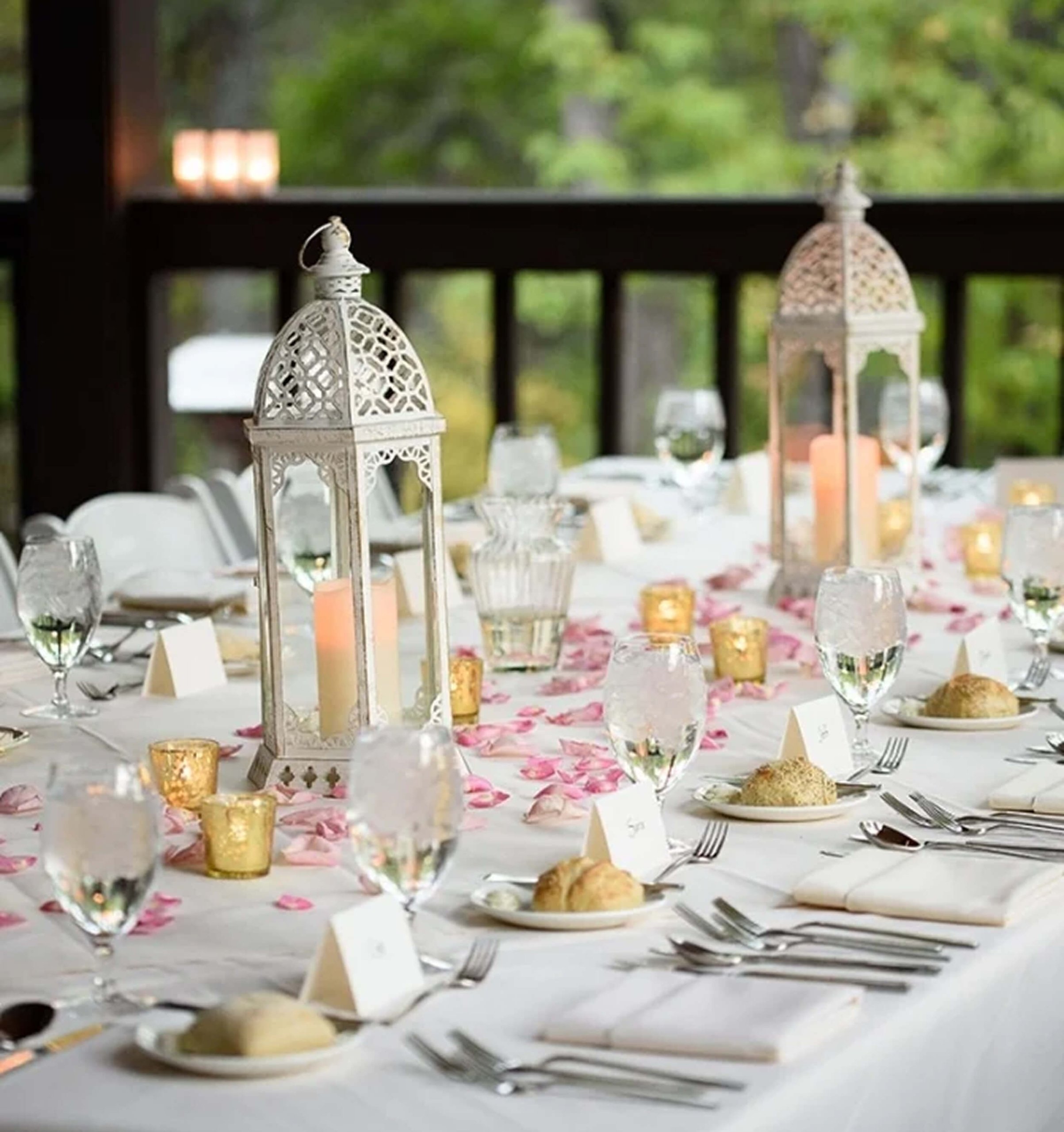 A long dining table set for a wedding, with white lanterns, wine glasses, and pink flower petals scattered on top.
