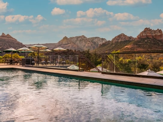View from the edge of a resort pool, looking over the water at the deck, umbrellas, and a panorama of Sedona's red rocks.