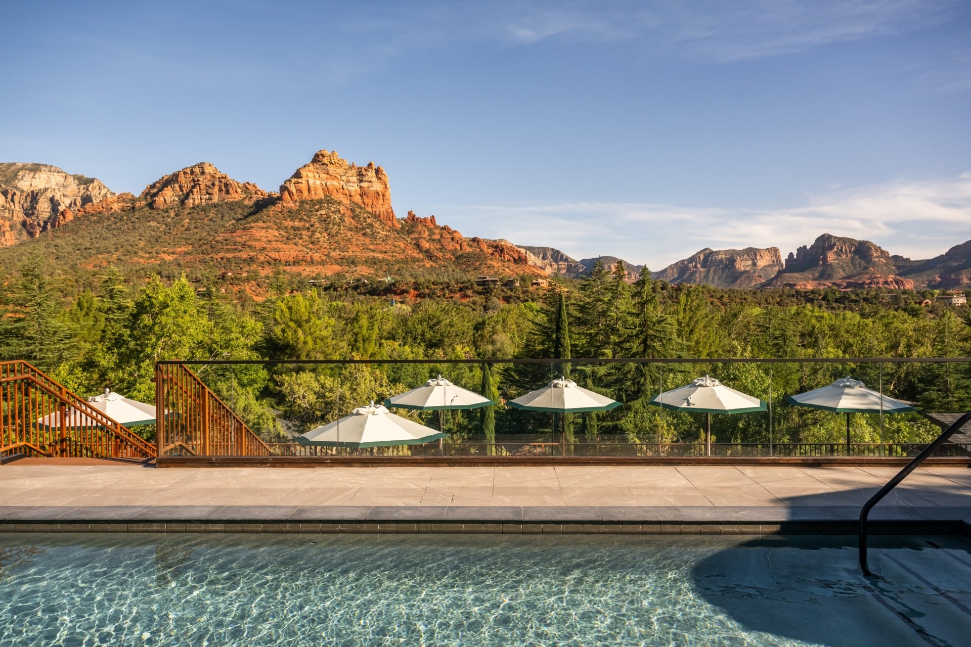 View from a swimming pool, looking past white umbrellas on the deck to a clear, panoramic view of Sedona's red rocks.