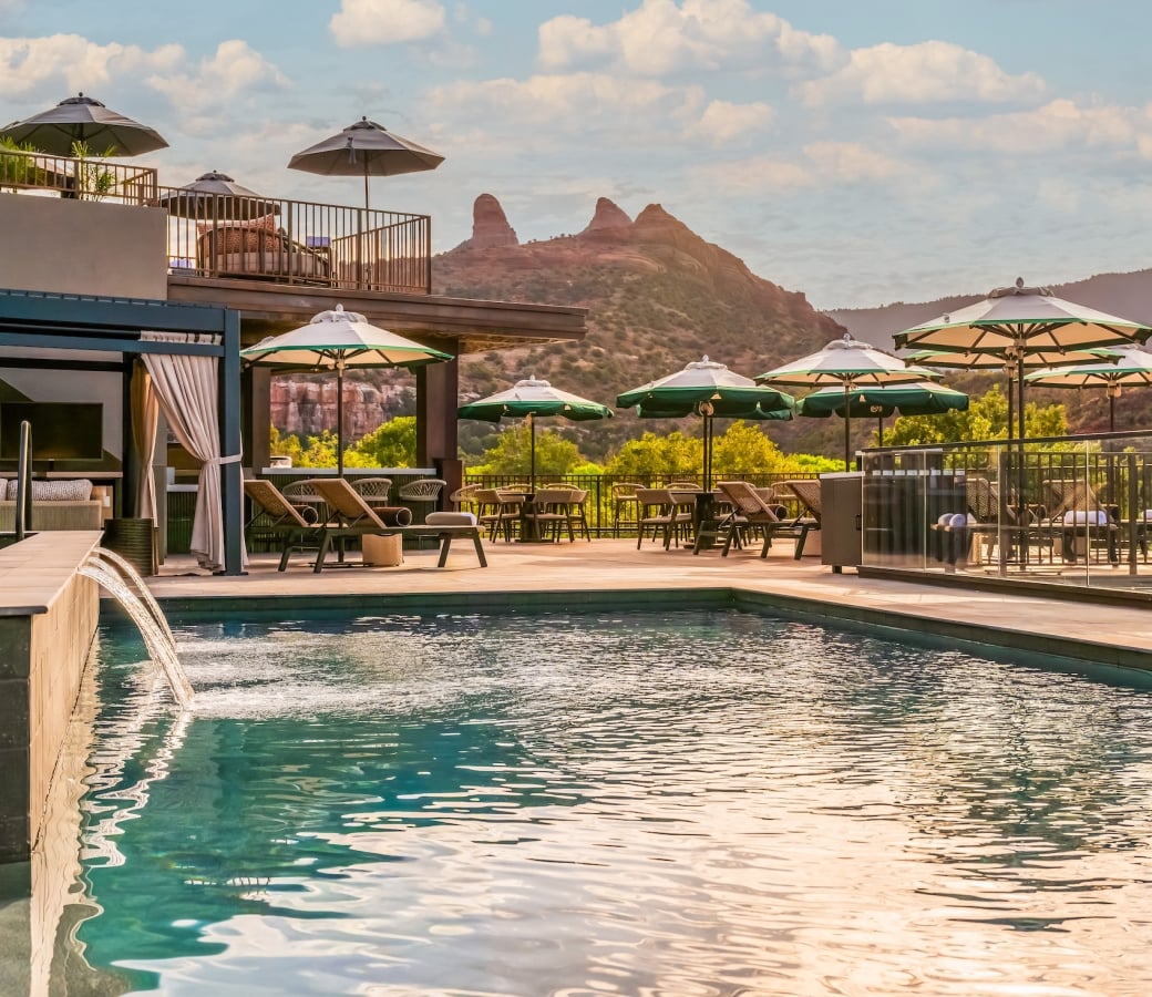 Resort pool with a water fountain, cabana, lounge chairs, and umbrellas, overlooking a terrace and Sedona's red rocks.