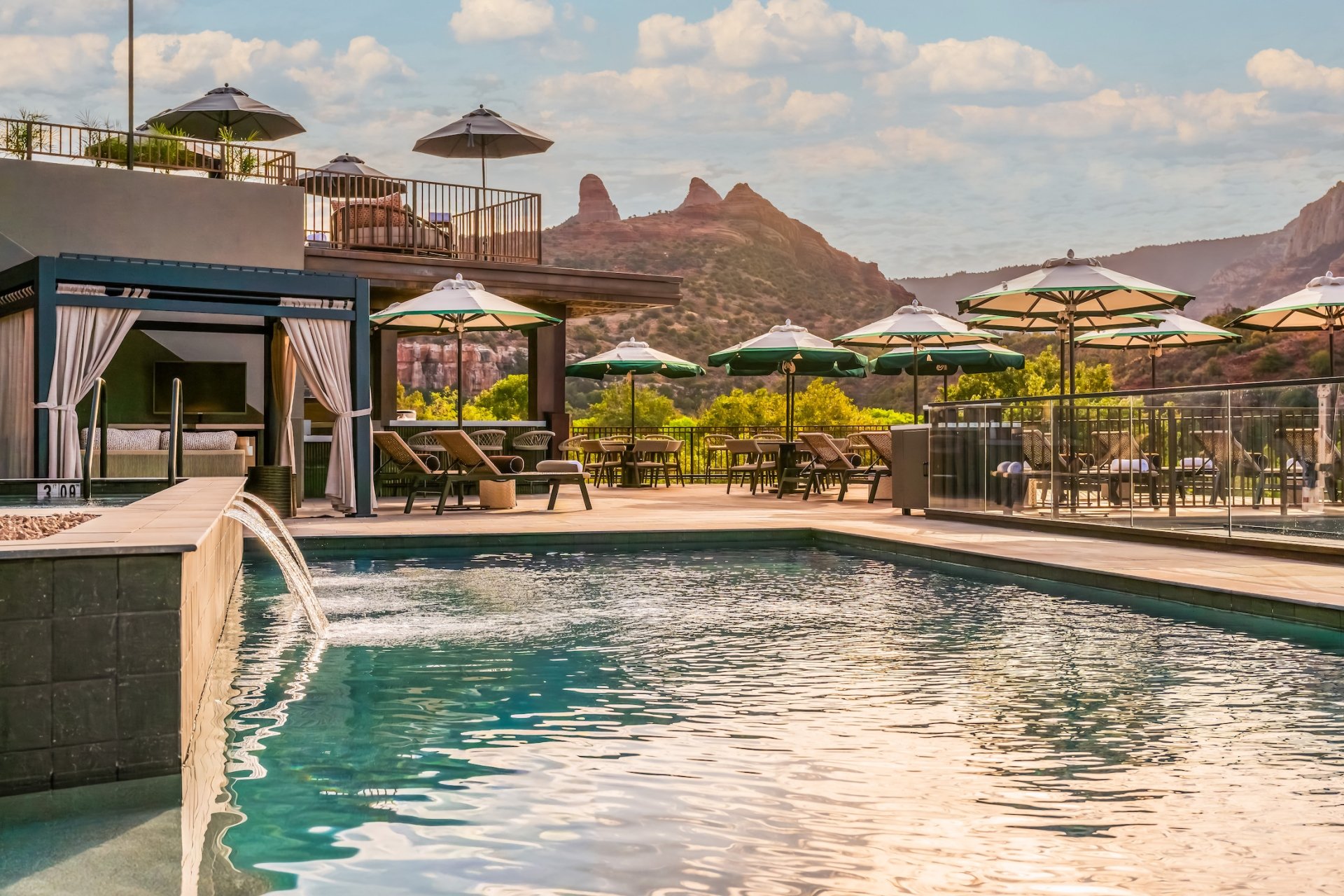 Resort pool with a water fountain, cabana, lounge chairs, and umbrellas, overlooking a terrace and Sedona's red rocks.