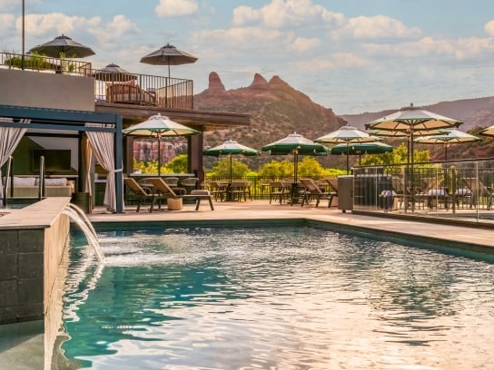 Resort pool with a water fountain, cabana, lounge chairs, and umbrellas, overlooking a terrace and Sedona's red rocks.