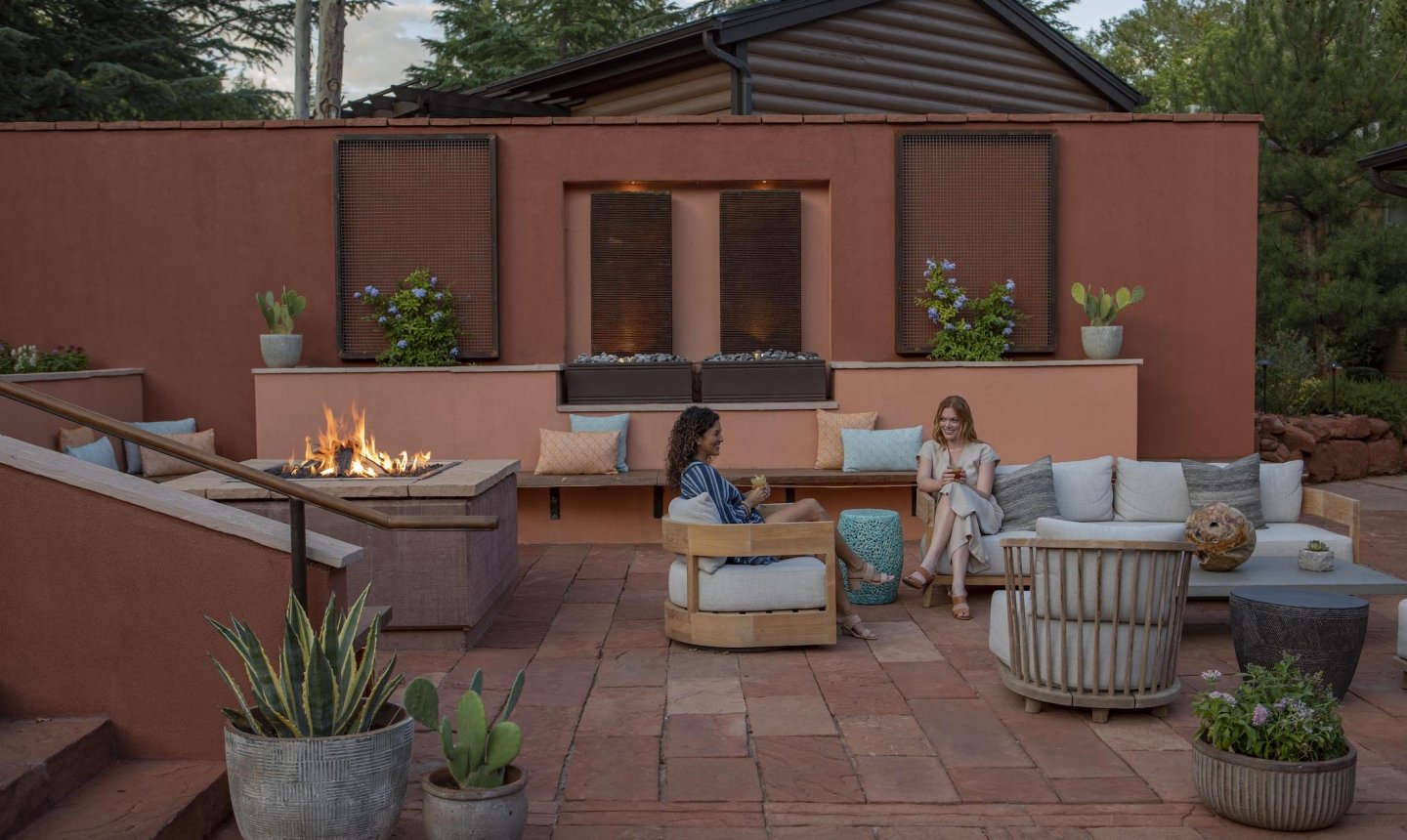 Two women talk while sitting in comfortable chairs on a red-tiled patio, gathered around a large, modern fire pit.