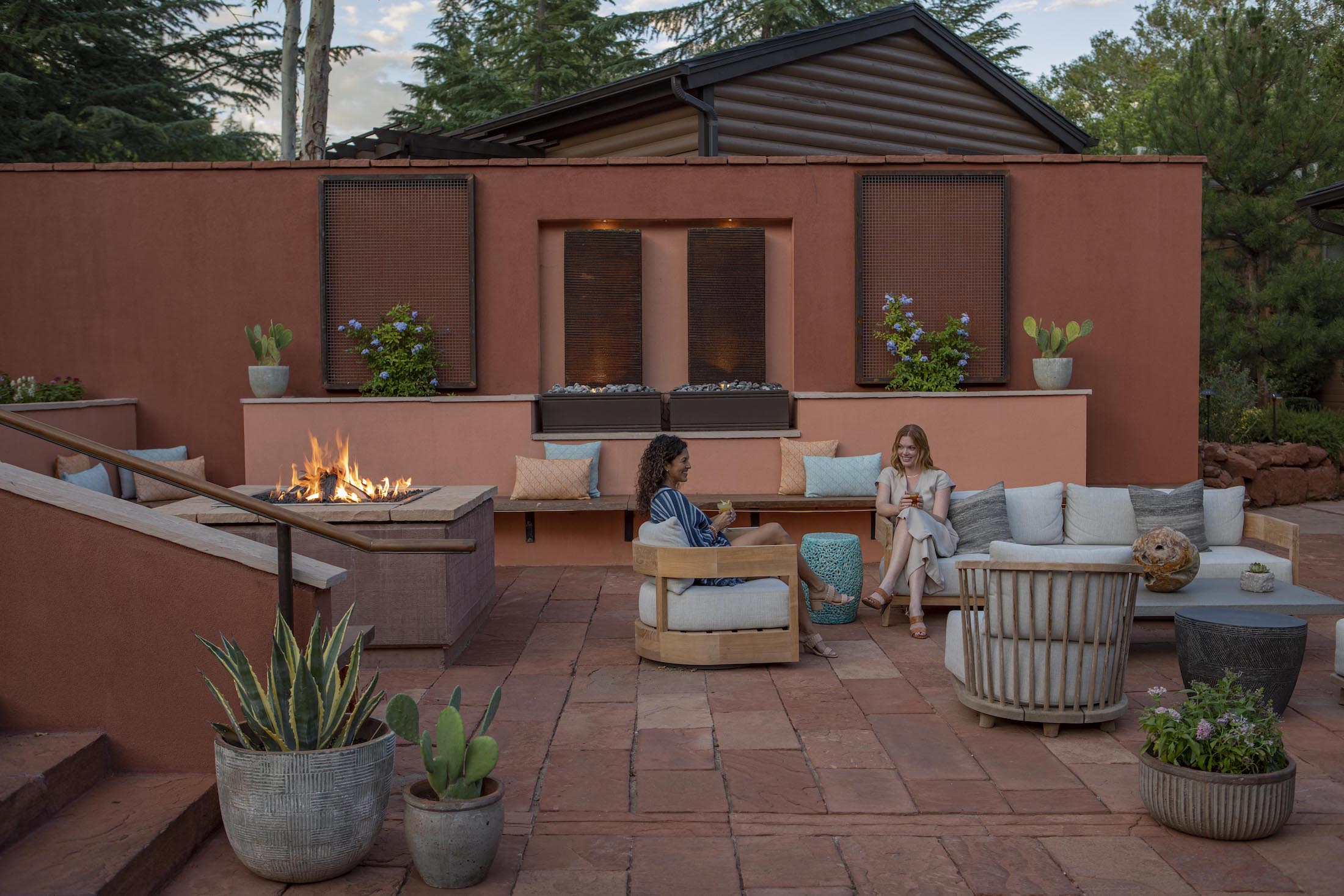 Two women talk while sitting in comfortable chairs on a red-tiled patio, gathered around a large, modern fire pit.
