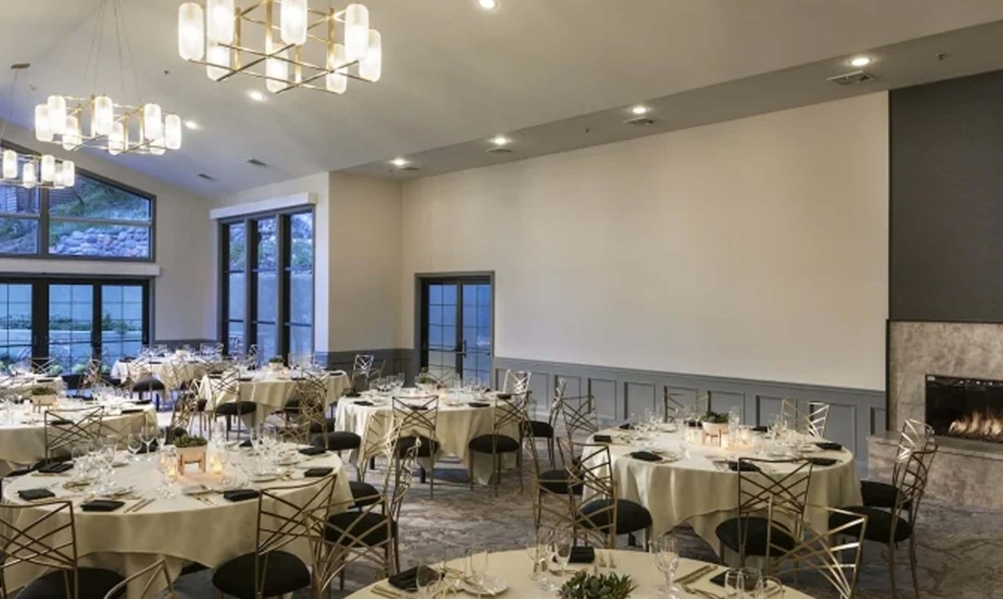 A modern ballroom set for a reception with round tables, gold chairs, and a fireplace.