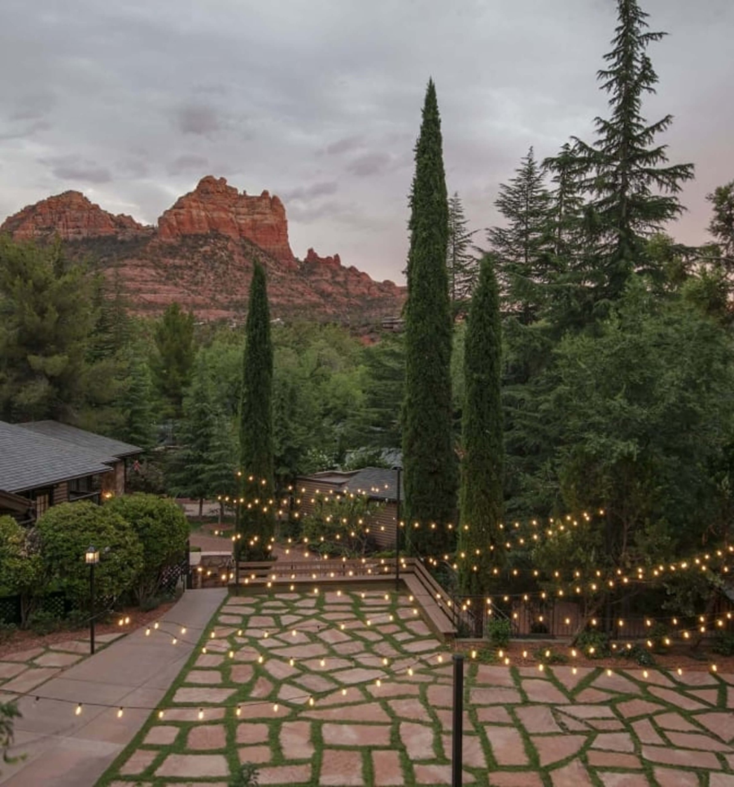 A stone patio with grass grout, illuminated by string lights, with cypress trees and Sedona's red rocks at dusk.