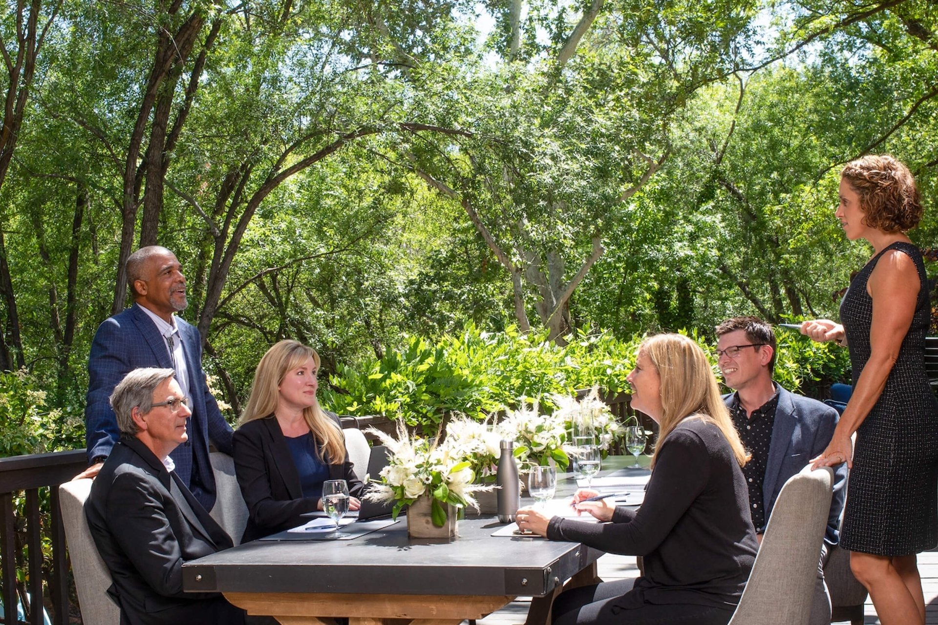 A group of professionals having an outdoor meeting at a dark table on a wooden deck, surrounded by lush green trees.