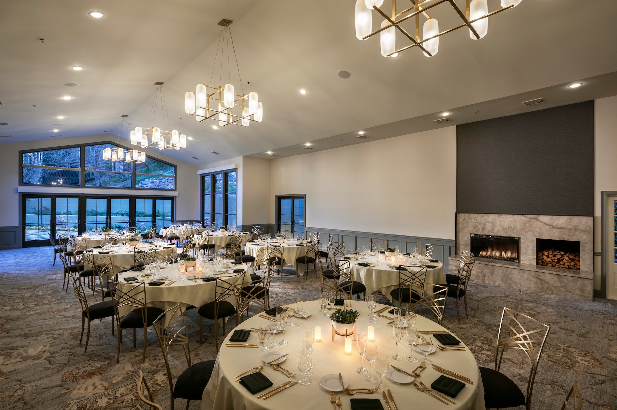 A modern ballroom set for a reception with round tables, gold chairs, modern chandeliers, and a large fireplace.