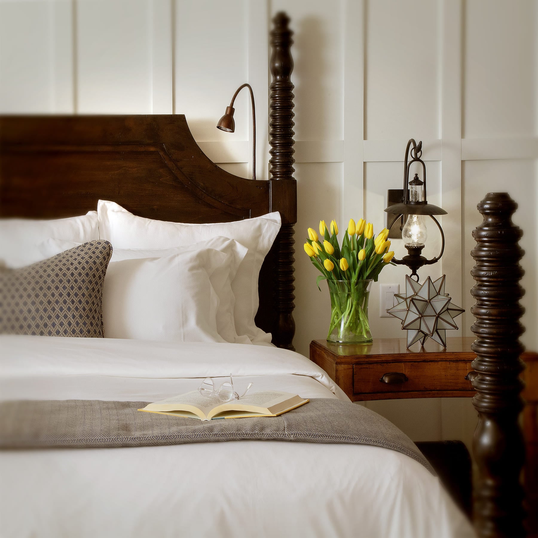 A close-up of a bed with white bedding, a dark wood headboard, and a nightstand with yellow tulips and a book.
