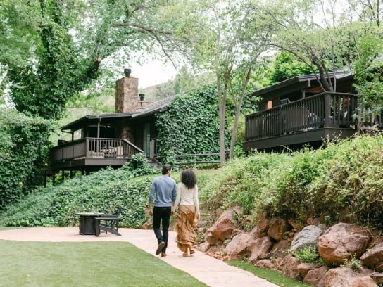A couple, seen from behind, walks on a garden path toward dark, ivy-covered hillside cottages at L'Auberge.