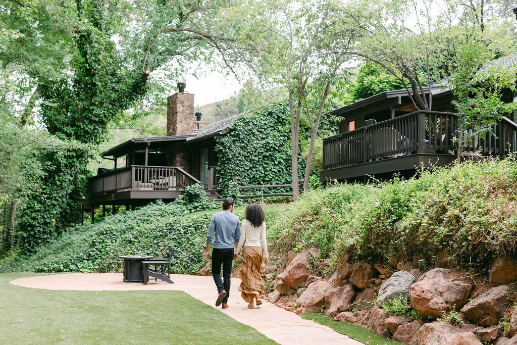 A couple, seen from behind, walks on a garden path toward dark, ivy-covered hillside cottages at L'Auberge.