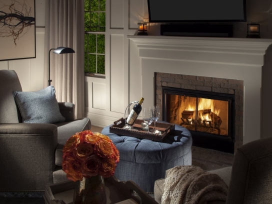 A cozy cottage room with a lit fireplace, an armchair, and a blue ottoman with a tray holding a wine bottle and glasses.