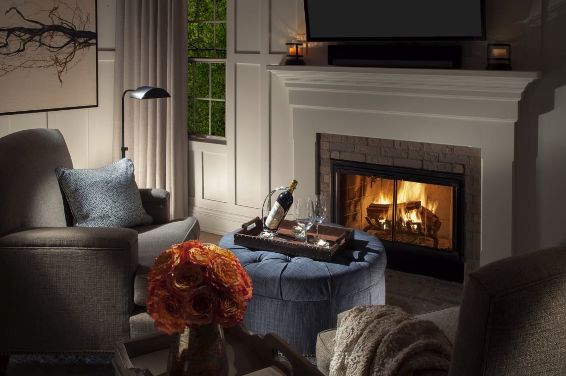 A cozy cottage room with a lit fireplace, an armchair, and a blue ottoman with a tray holding a wine bottle and glasses.