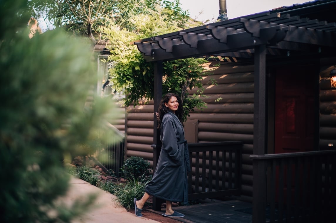 A woman in a dark gray bathrobe smiles as she walks on the wooden deck of a log cabin, surrounded by greenery.