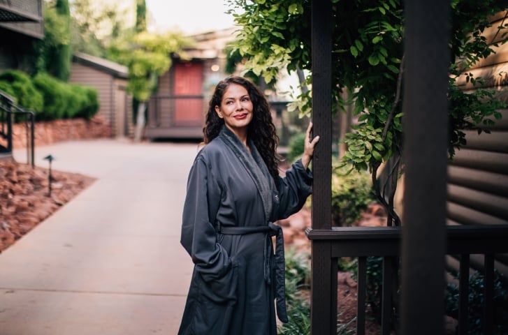 A smiling woman with long curly hair, wearing a gray robe, stands on a garden path next to a log cabin.