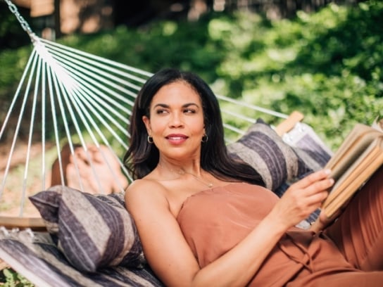 A woman in a brown top relaxes in a hammock with striped pillows, holding a book in a sunny garden.