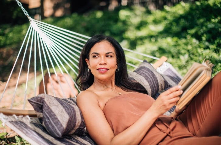 A woman in a brown top relaxes in a hammock with striped pillows, holding a book in a sunny garden.