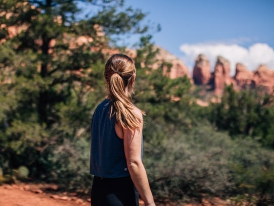 A woman with a ponytail, seen from behind, stands on a trail looking out at a scenic view of Sedona's red rocks.