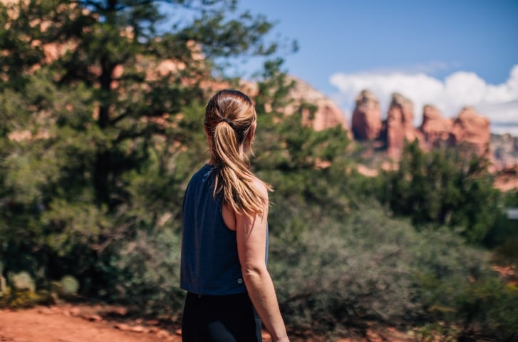 A woman with a ponytail, seen from behind, stands on a trail looking out at a scenic view of Sedona's red rocks.