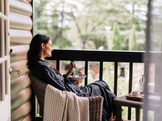 A woman in a robe relaxes with a bowl of fruit on a wicker chair on a private, wooded cottage balcony.