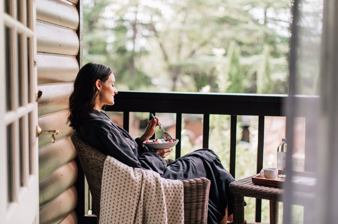 A woman in a robe relaxes with a bowl of fruit on a wicker chair on a private, wooded cottage balcony.