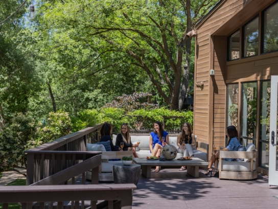 A group of women relaxes and drinks wine on a comfortable outdoor lounge set on a wooden deck, surrounded by lush trees.