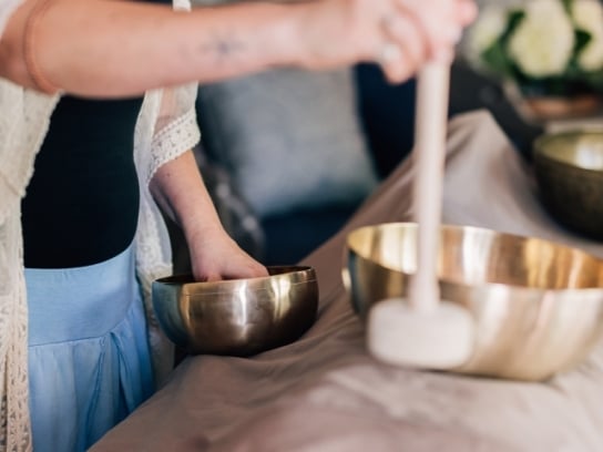 A close-up of a sound healing session, showing a practitioner's hands playing two large, golden singing bowls.