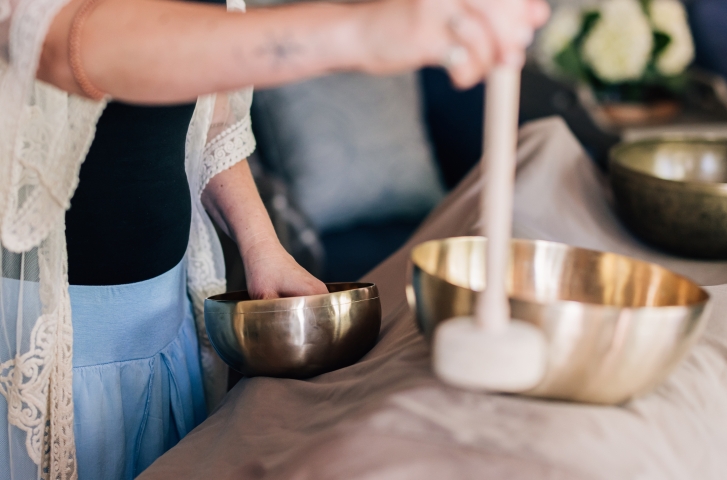 Close view shows a therapist in a lace kimono and pale blue skirt beside a table draped in taupe linen, one hand in a brass singing bowl and a padded striker over another bowl.