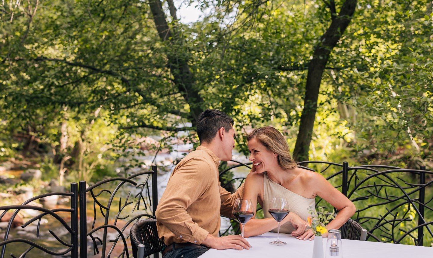 A smiling couple drinks red wine at a white-clothed table on a patio, with the lush greenery of Oak Creek behind them.
