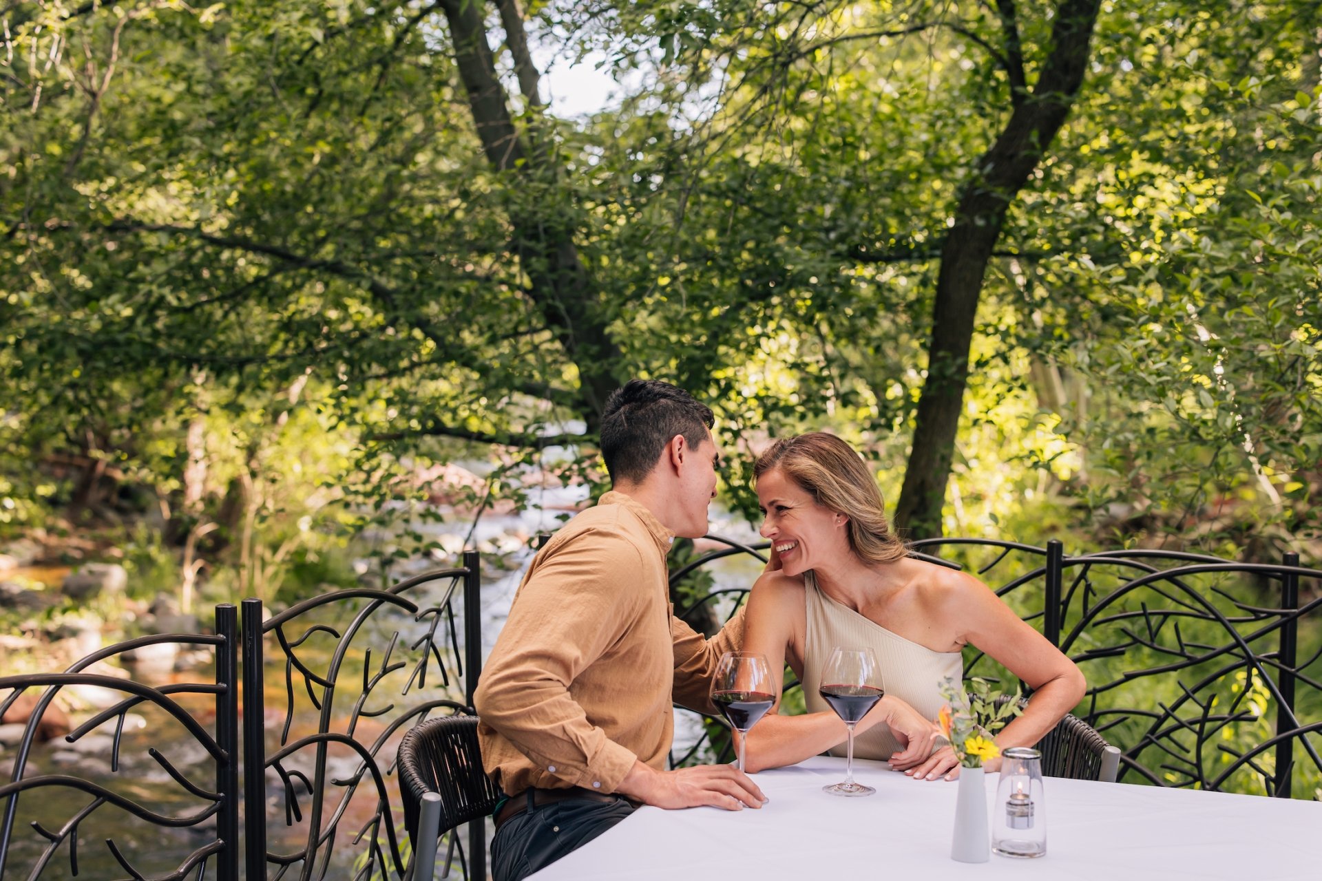 A smiling couple drinks red wine at a white-clothed table on a patio, with the lush greenery of Oak Creek behind them.