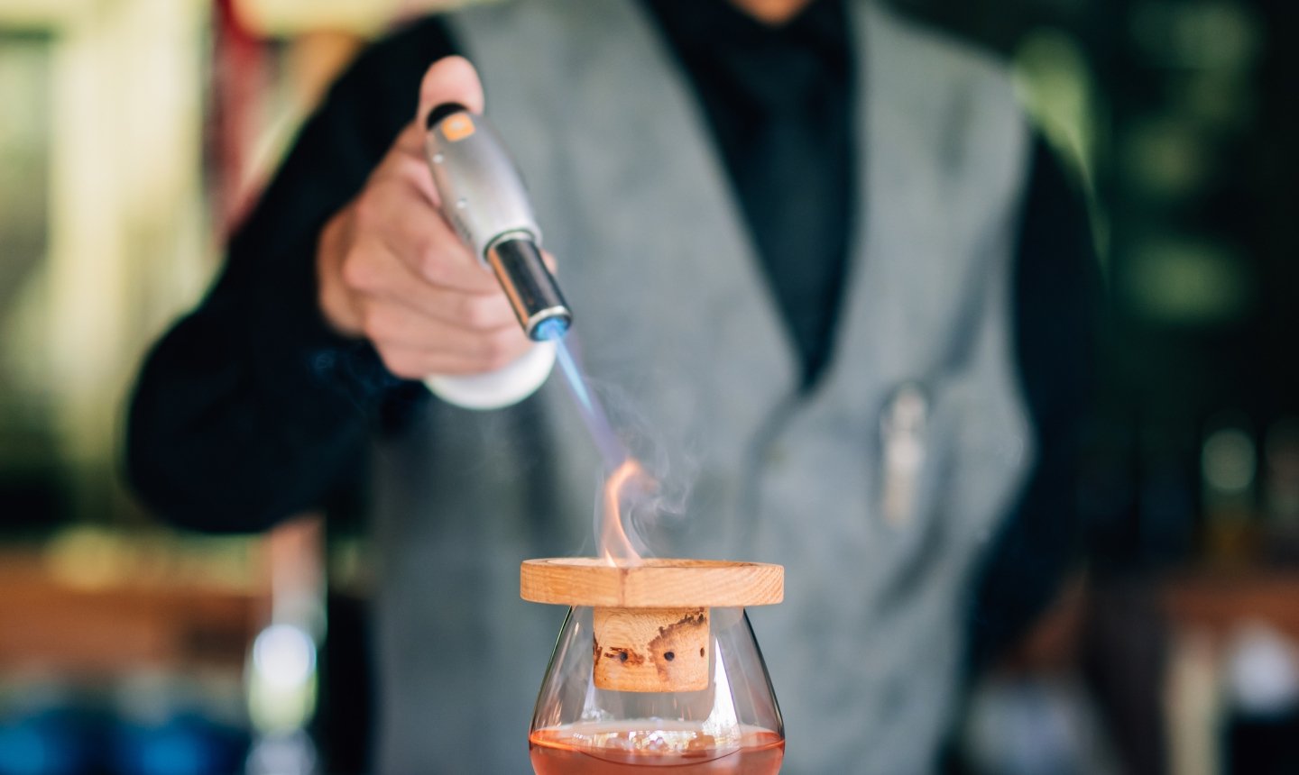 A bartender in a vest uses a culinary torch to light a wood chip, smoking a dark-colored cocktail in a snifter glass.
