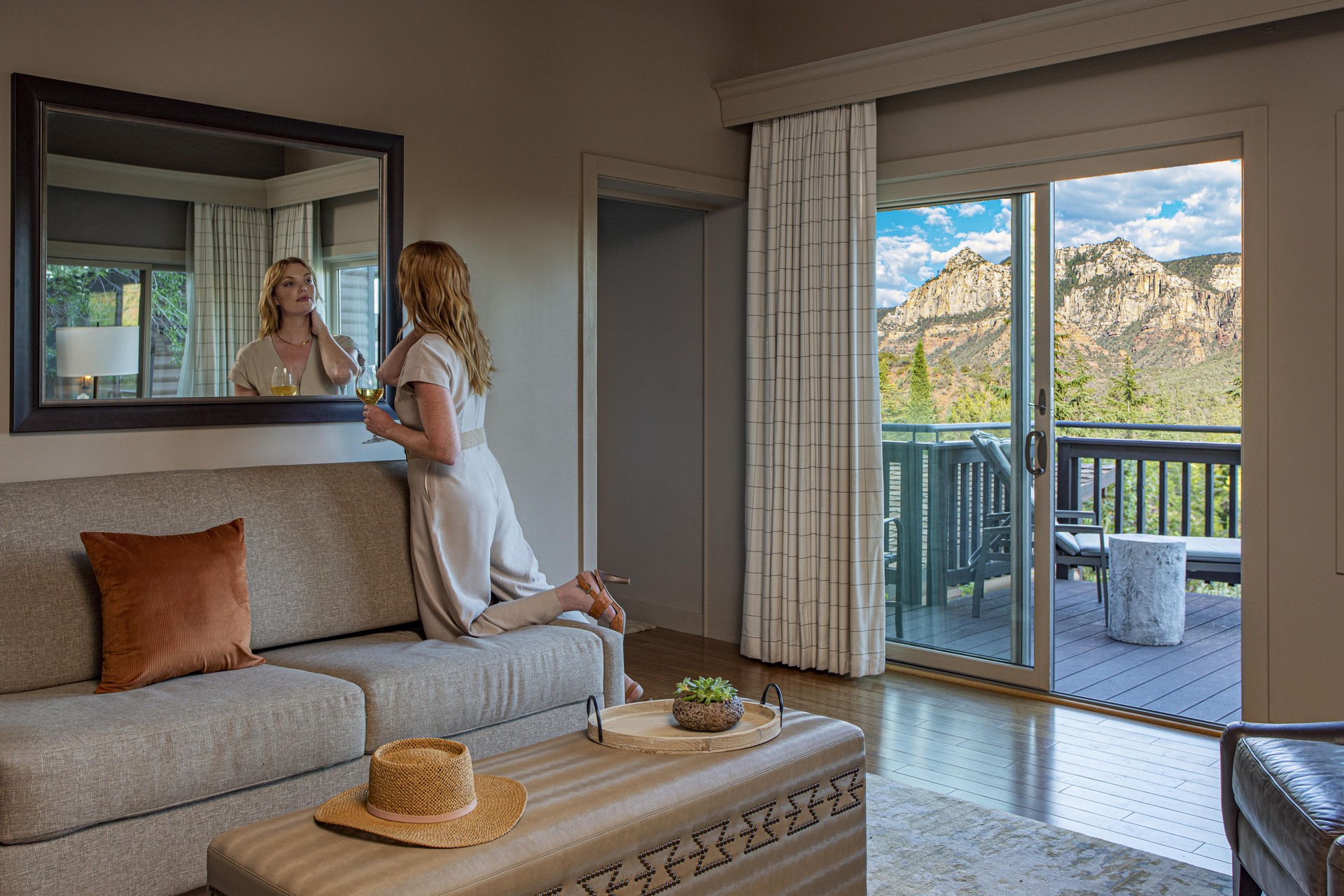 A woman sits on a couch in a living room with a sliding glass door leading to a balcony with a mountain view.