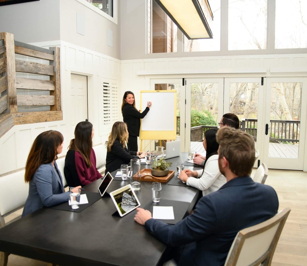 A woman presents on a whiteboard to a group of colleagues seated at a long table in a bright, modern meeting room.