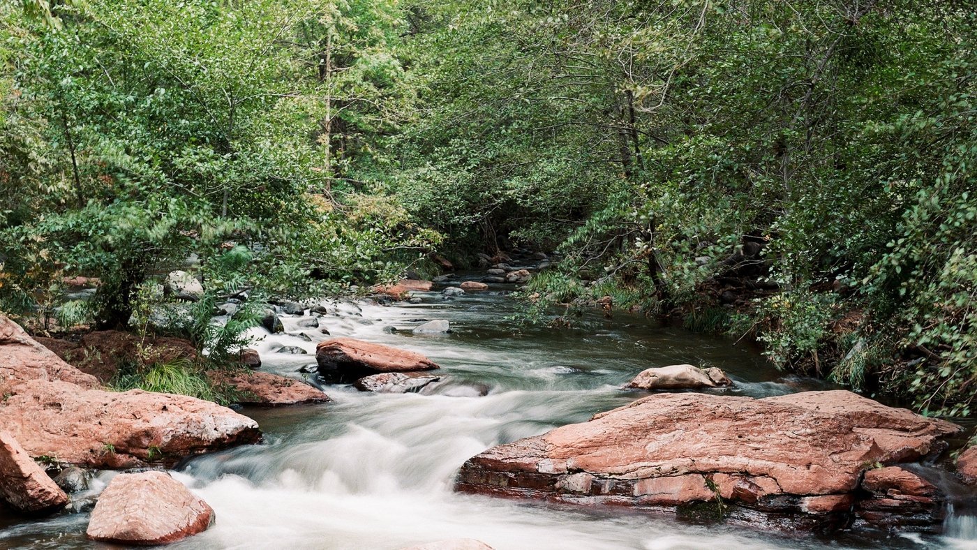 A long-exposure photo of Oak Creek, showing silky white water flowing over red rocks in a lush, green forest.