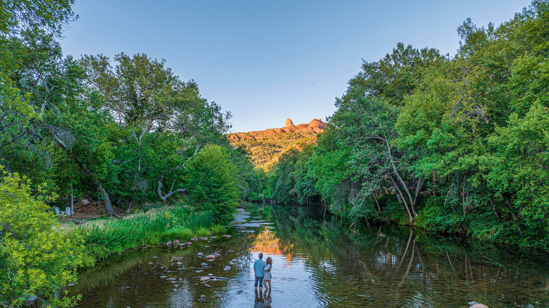 A couple stands in the shallow water of Oak Creek, surrounded by lush green trees that reflect on the water's surface.