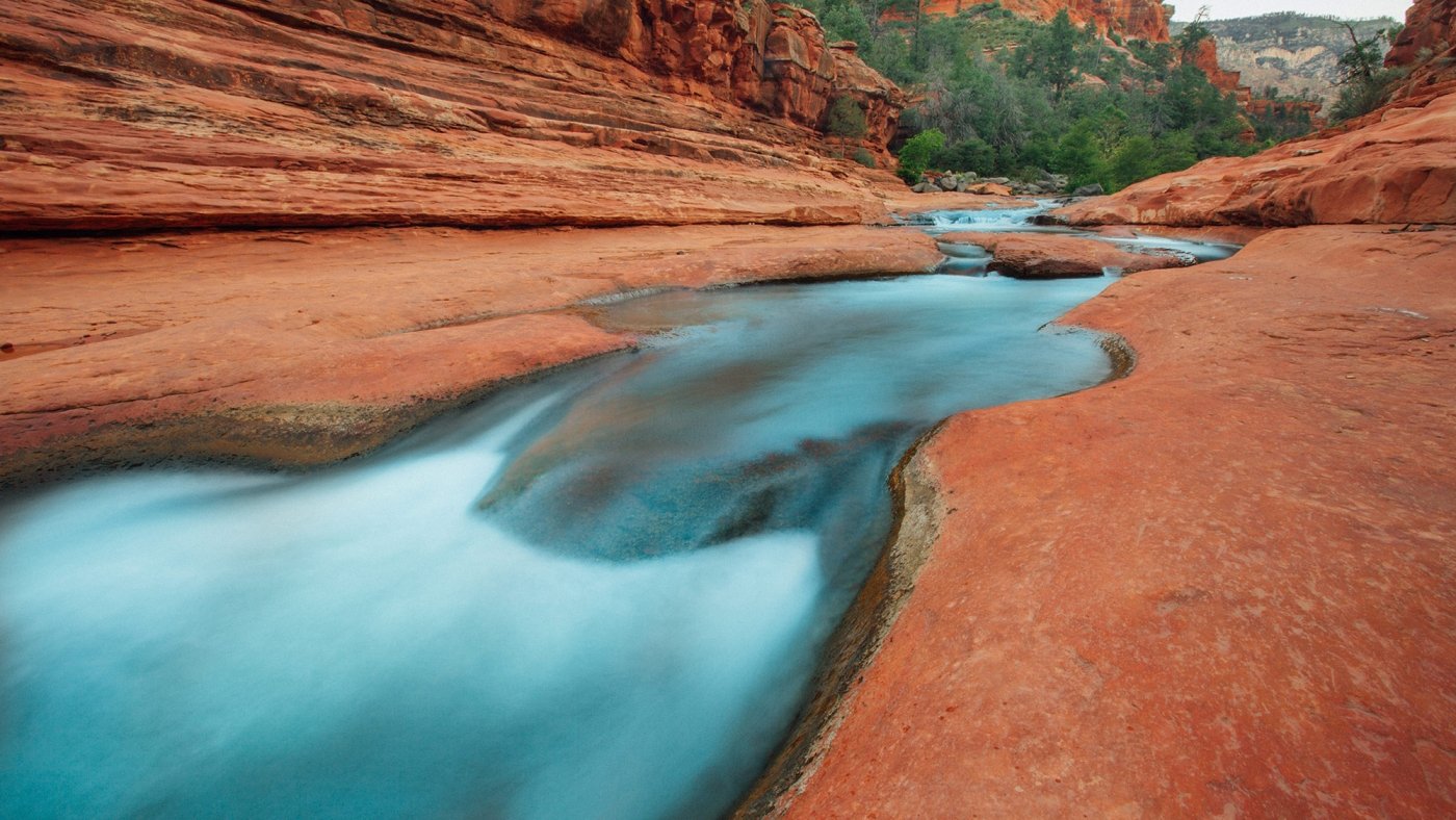 A long-exposure shot of silky, blurred water flowing through the smooth, carved red rock canyon of Slide Rock.