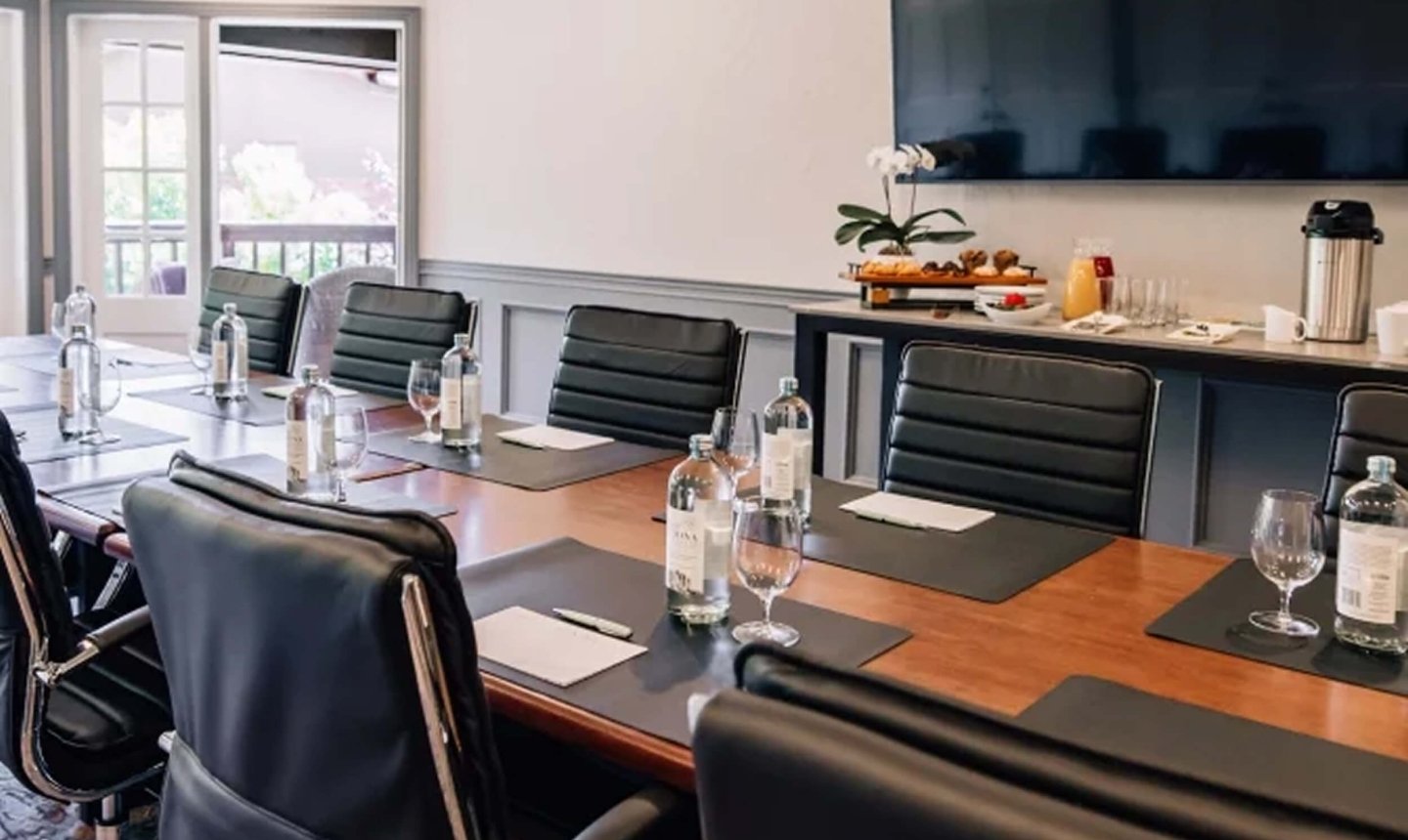 A professional boardroom with a long wooden table, black leather chairs, a large screen, and a refreshment station.
