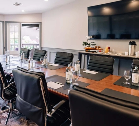 A professional boardroom with a long wooden table, black leather chairs, a large screen, and a refreshment station.