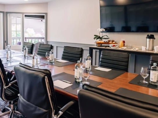 A professional boardroom with a long wooden table, black leather chairs, a large screen, and a refreshment station.