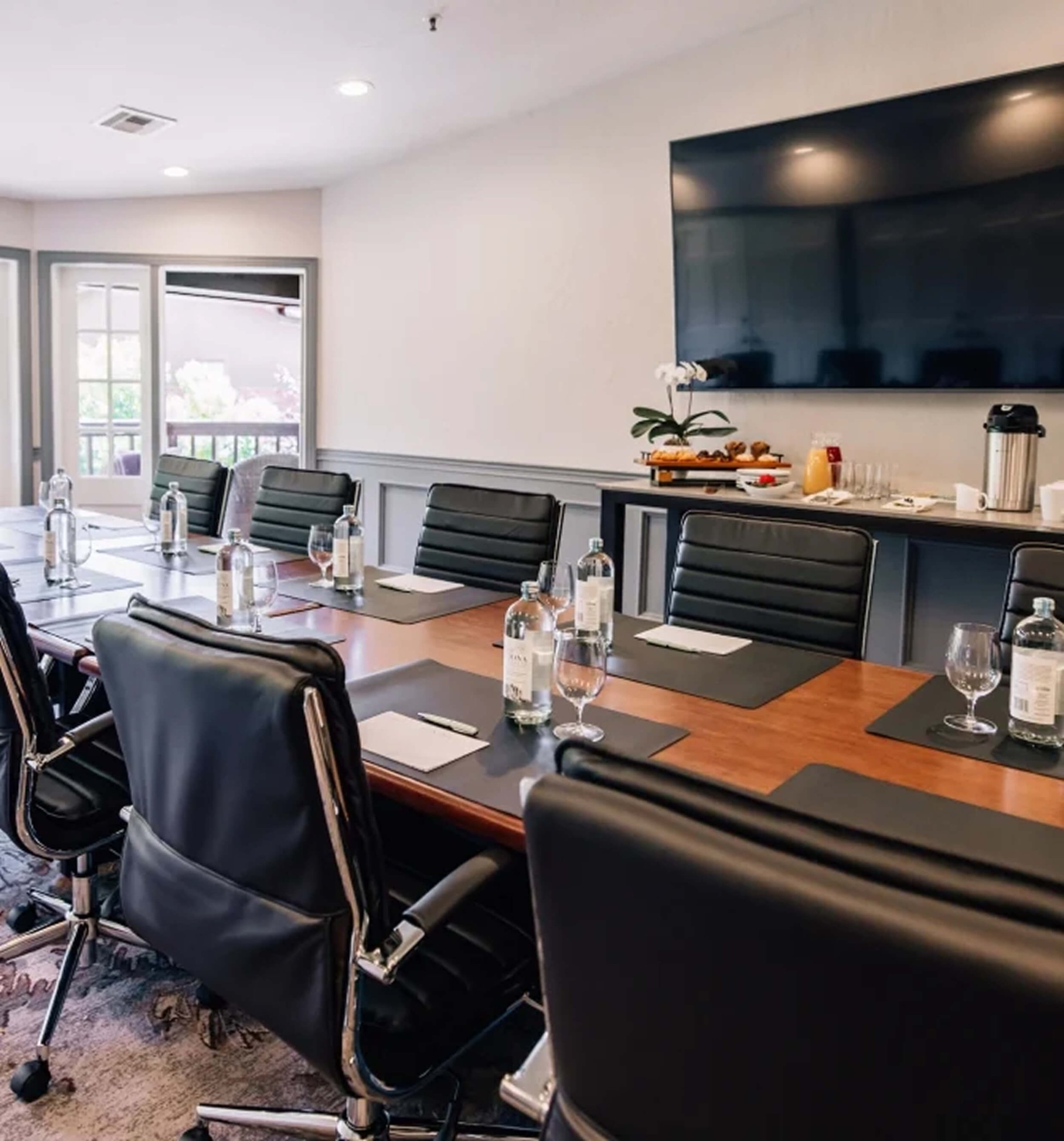 A professional boardroom with a long wooden table, black leather chairs, a large screen, and a refreshment station.