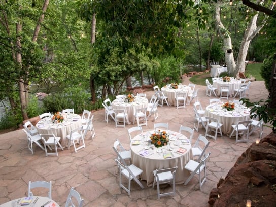 An outdoor wedding reception on a stone patio with round tables and white chairs, set among trees next to a creek.