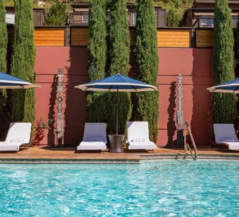 White lounge chairs and blue-striped umbrellas line a sparkling blue pool, set against a red wall and tall cypress trees.